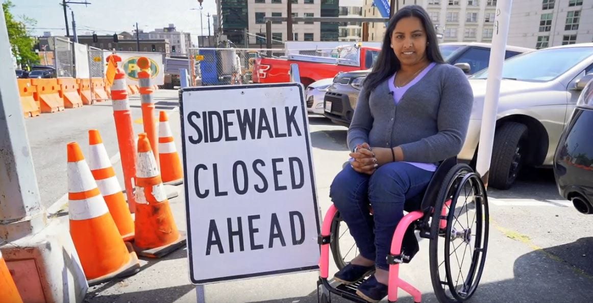 Anita sits in front of a Sidewalk Closed sign on a sidewalk in Downtown Seattle.