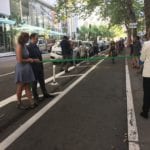Sam Zimbabwe, Vicky Clark, and Mayor Jenny Durkan hold the ribbon as a person biking approaches. Celebrating the opening of the bike lane!