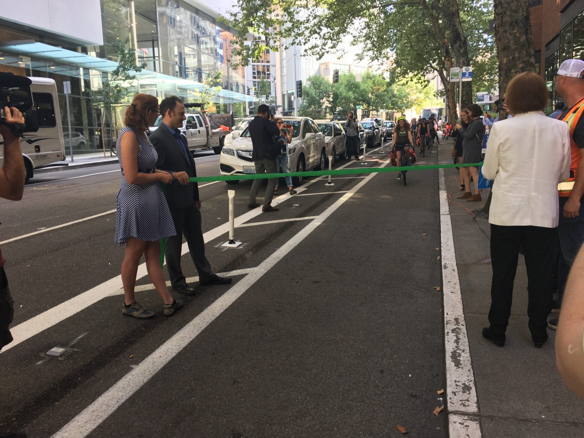 Sam Zimbabwe, Vicky Clark, and Mayor Jenny Durkan hold the ribbon as a person biking approaches. Celebrating the opening of the bike lane!