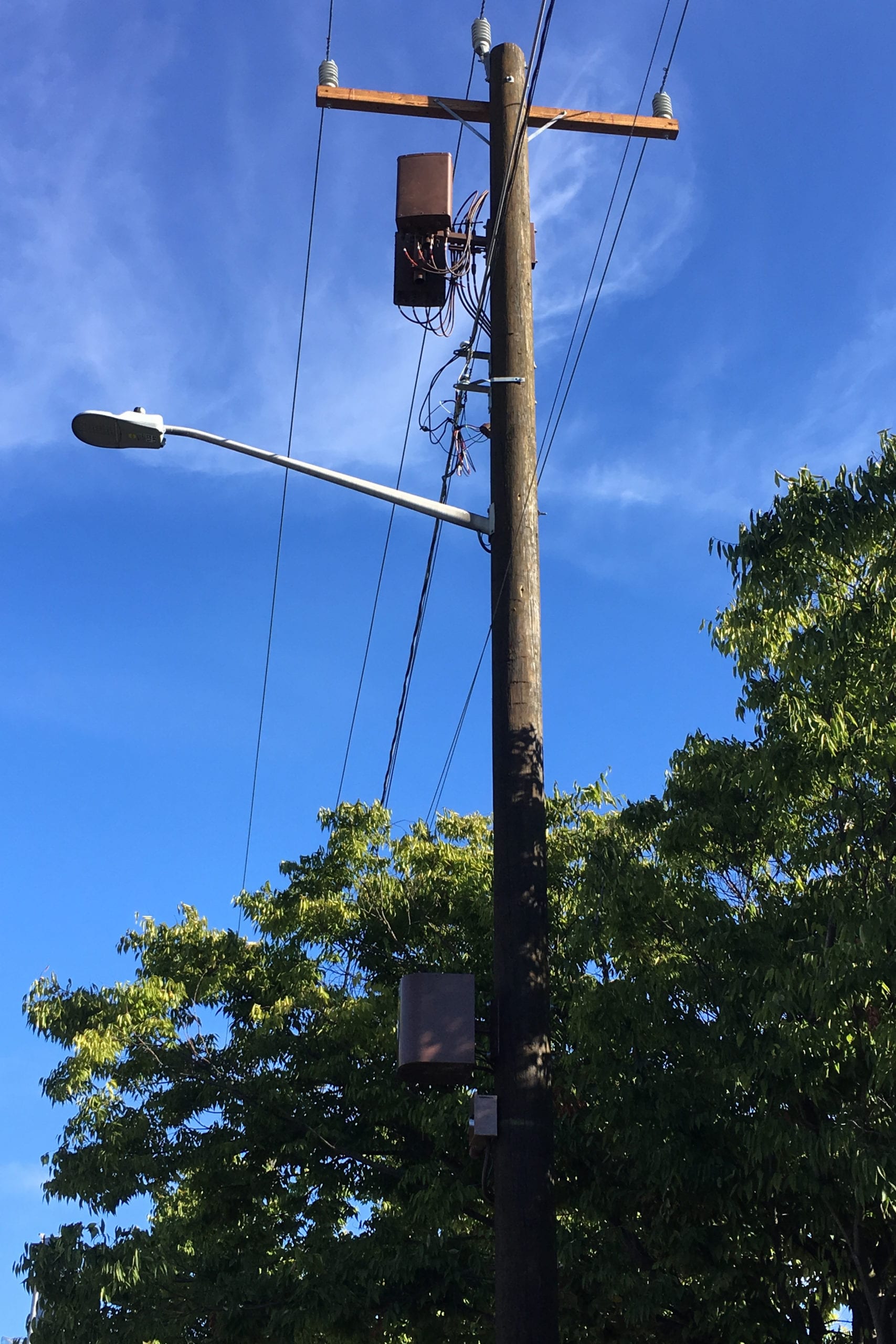 A wooden utility pole with a streetlight, overhead power lines, and a side-mounted small wireless facility between the power lines and the streetlight