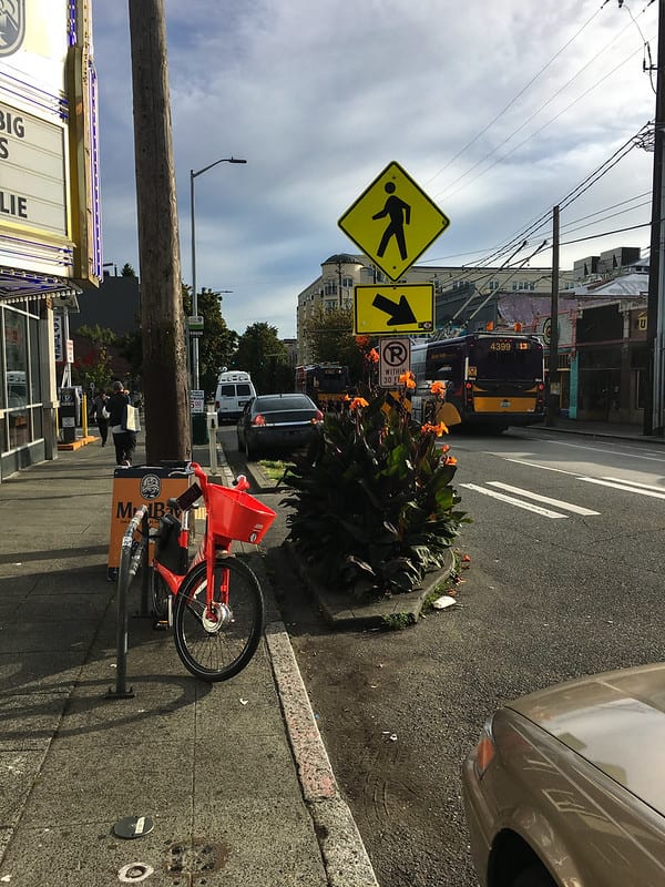 Red bike-share bike resting against a bike rack on a sidewalk, near a crosswalk sign with orange flowers growing next to it.