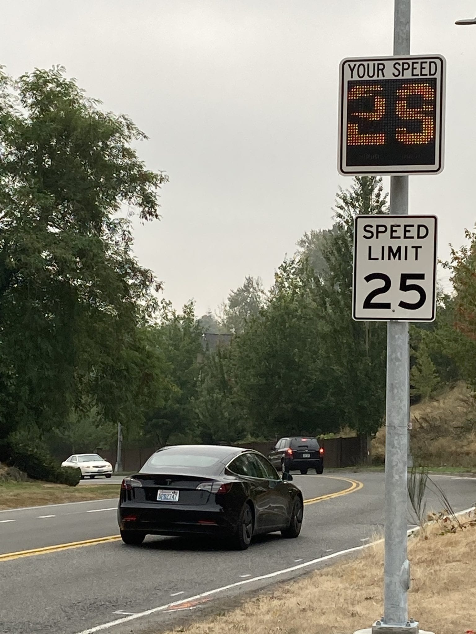 Several cars going by a speed monitoring device showing a speed limit of 25 mph.