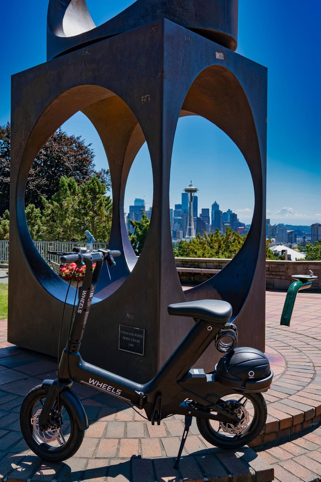 Wheels scooter in front of a sculpture on a sunny day in Seattle. Downtown and the Space Needle can be seen in the background.