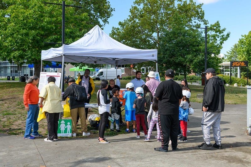 Group of adults and children at a pop-up outreach event at Rainier Ave S and S Henderson St. Event is taking place on pavement near a grassy area on a sunny day. Trees are shown in background.