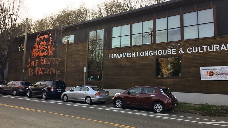 The Duwamish Longhouse and Cultural Center shown from the outside, viewed from West Marginal Way. Four cars are parked in front. Photo Credit: SDOT Flickr. 