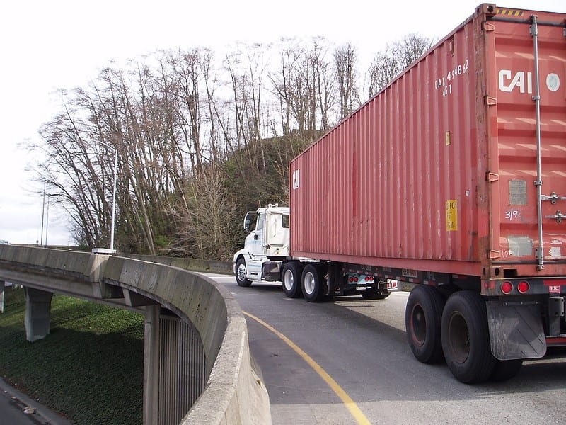 A semi-truck with a white cab and red freight area is shown driving up a ramp.