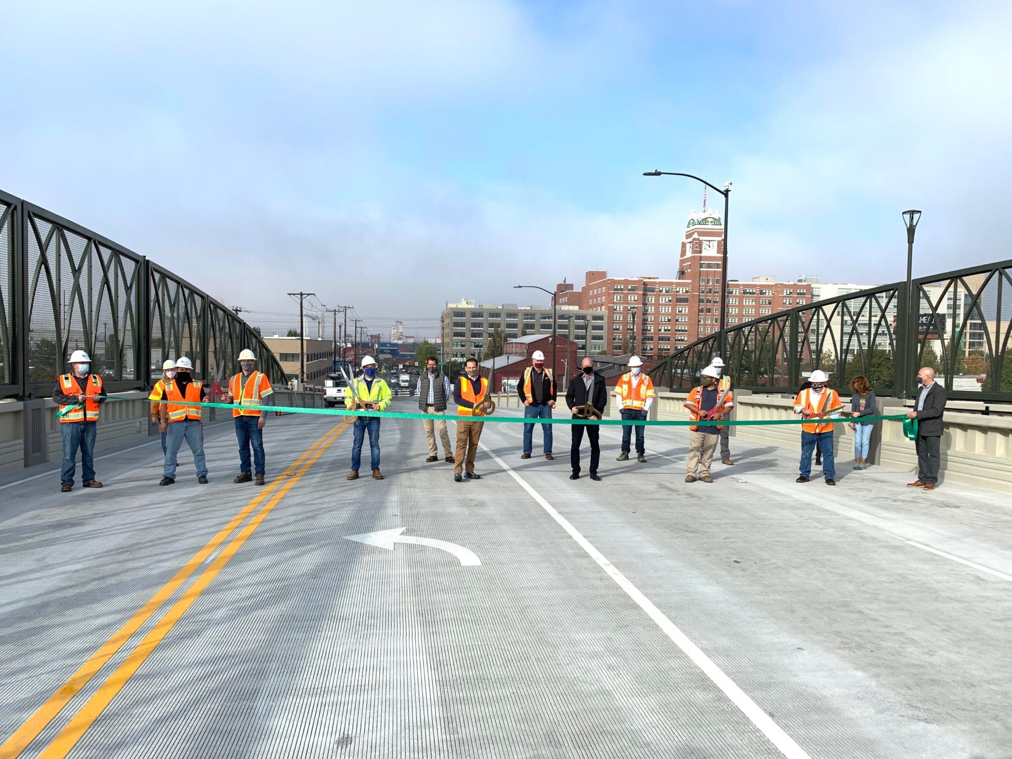 Group of people in construction vests and hard hats holding a green ribbon across the Lander St Bridge. One person holds large scissors and is preparing to cut the ribbon.