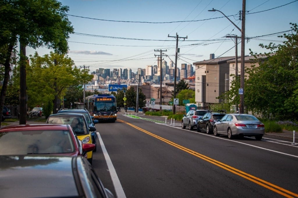 A bus is shown coming up the SW Avalon Way hill on a newly repaved road. Downtown Seattle is seen in the background.