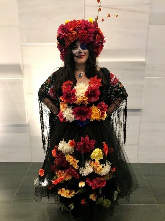Sonia dressed as Catrina, the “Lady of the Dead” - one of the most widely recognized symbols of Dia de Muertos.  She is wearing colorful black, yellow, orange, and red attire, a floral headdress with the same colors, and white face paint to evoke an image of a beautiful skull.