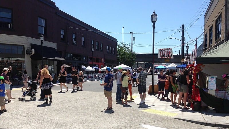 A street festival in Georgetown during pre-pandemic days. People are seen milling about near tents and umbrellas. It looks like a warm summer day. A primary focus of Reconnect West Seattle is to support those communities, like Georgetown, which are deeply impacted by the bridge closure and related detour routes. 