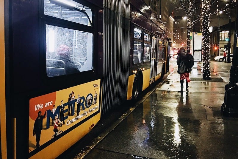 Person walking on the sidewalk next to a bus in the rain. Holiday lights are shown wrapped around trees on street.