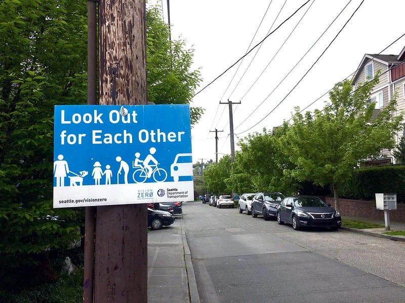 Vision Zero sign on a wooden pole on a neighborhood street. Sign is blue with white writing and icons of people. One person is walking a dog, two children are holding hands, one person is walking with a cane, and one person is riding a bike with a child on the back of the bike, and there is a car. The writing says, "look out for each other."