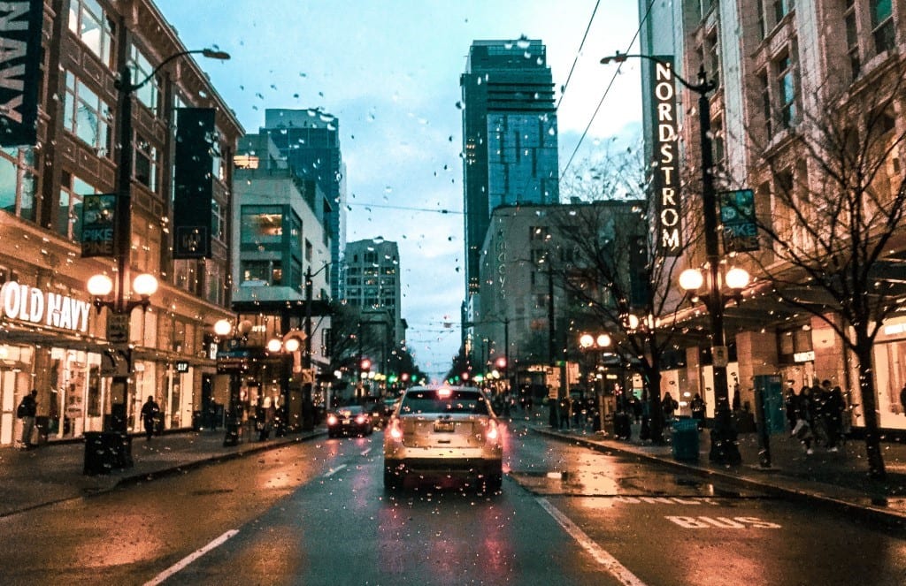 Photo taken from inside a vehicle looking out the windshield. Vehicle is behind another vehicle. It is late afternoon and rainy. The vehicle is driving in downtown Seattle past Old Navy and Nordstrom.