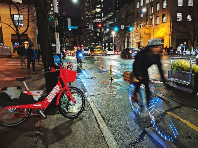 Person biking in a bike lane with blue lights on wheels. It is nighttime. There is a red jump bike to the person's right, on the sidewalk.