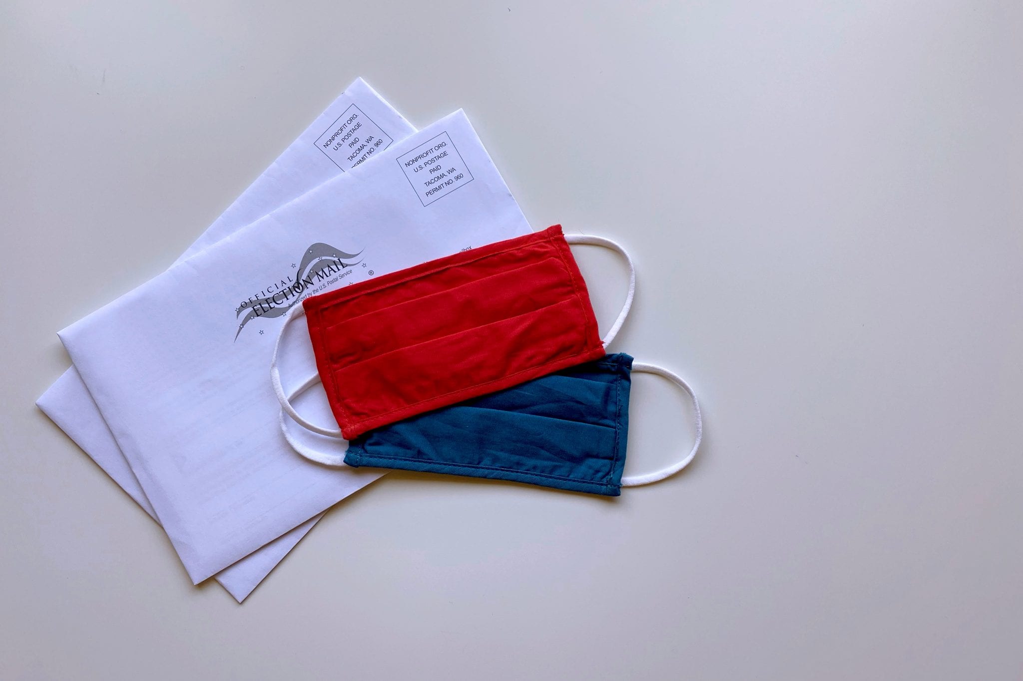 Red mask and blue mask on top of two election mail in ballots in envelopes on a white background.