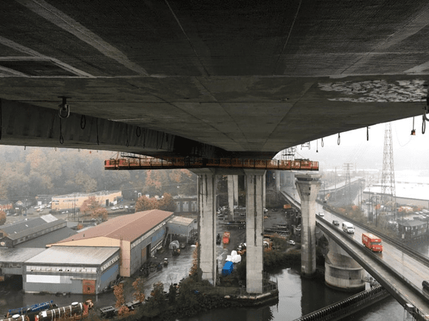 Temporary work platforms under the West Seattle High Rise Bridge. Platforms are orange and span the width of the bridge, and are suspended underneath is. Photo is taken looking at the full platform, looking west. The Low Bridge with vehicles can be seen in the lower right corner of the image. It is a rainy day.