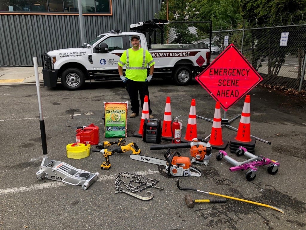Person standing behind all the materials contained in an SDOT response truck, inlcuding a pink "Emergency scene ahead" sign, traffic cones, emergency flashers, and an array of other materials.