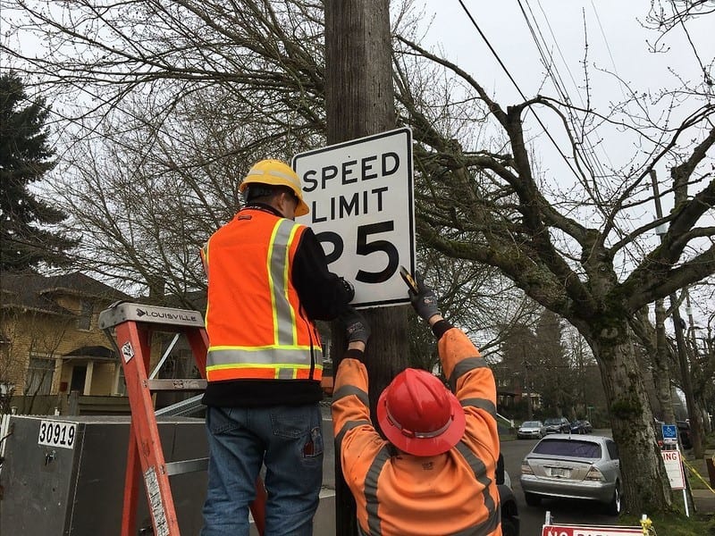 Two crew members hanging a 25 MPG speed limit sign on a wooden post using a ladder.