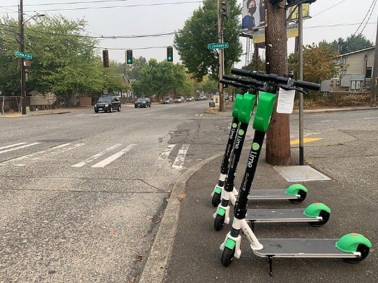 Three lime scooters on a sidewalk in Seattle next to an intersection. Two vehicles are approaching the intersection.