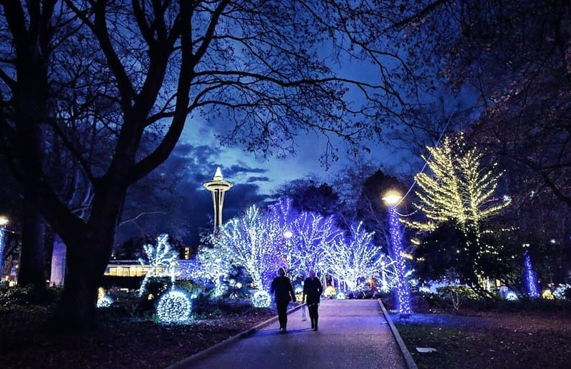 People walking at night through Denny Park. There are blue and yellow holiday lights in the park, and the Space Needle can be seen in the background.