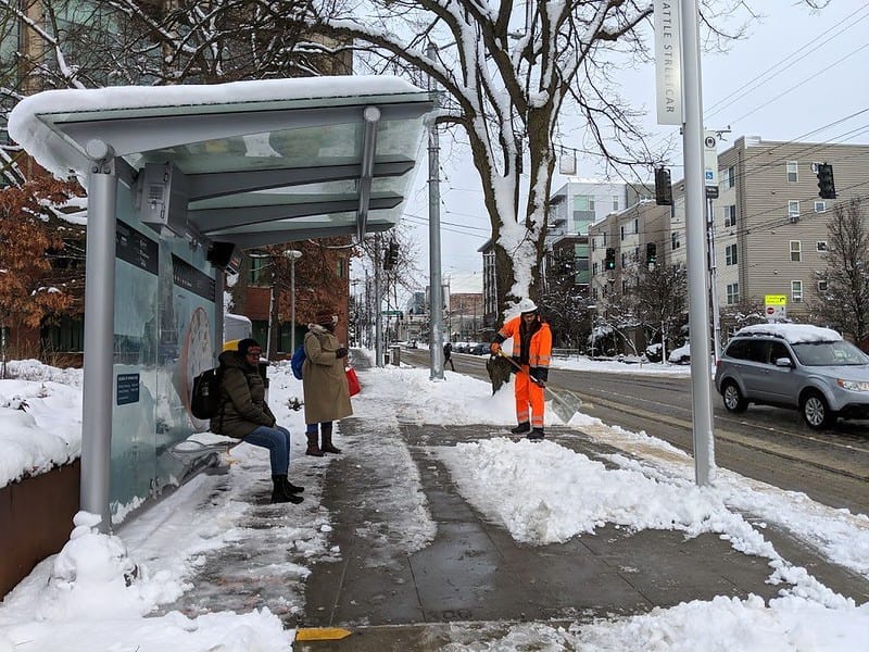 People waiting at a streetcar shelter while a crew member shovels snow nearby. Snow is on the ground around the shelter.