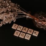 Scrabble tiles that spell out the words Thank You. Above the tiles rests foliage in a vase.