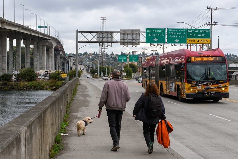 Two people walking away from the camera with a small dog. The people are walking across the West Seattle Low Bridge. To the peoples' right, a Rapid Ride bus is heading towards the camera.