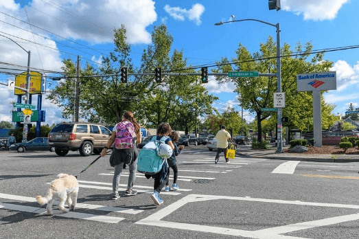 Children, adults, and a dog crossing S Henderson St. along Rainier Ave S on a sunny day.