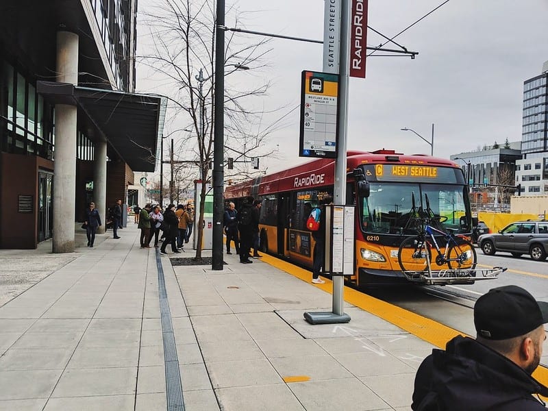 Downtown Seattle viewed from the sidewalk. A RapidRide C-Line bus is stopped at a bus stop. People are seen waiting for the bus and boarding the bus.