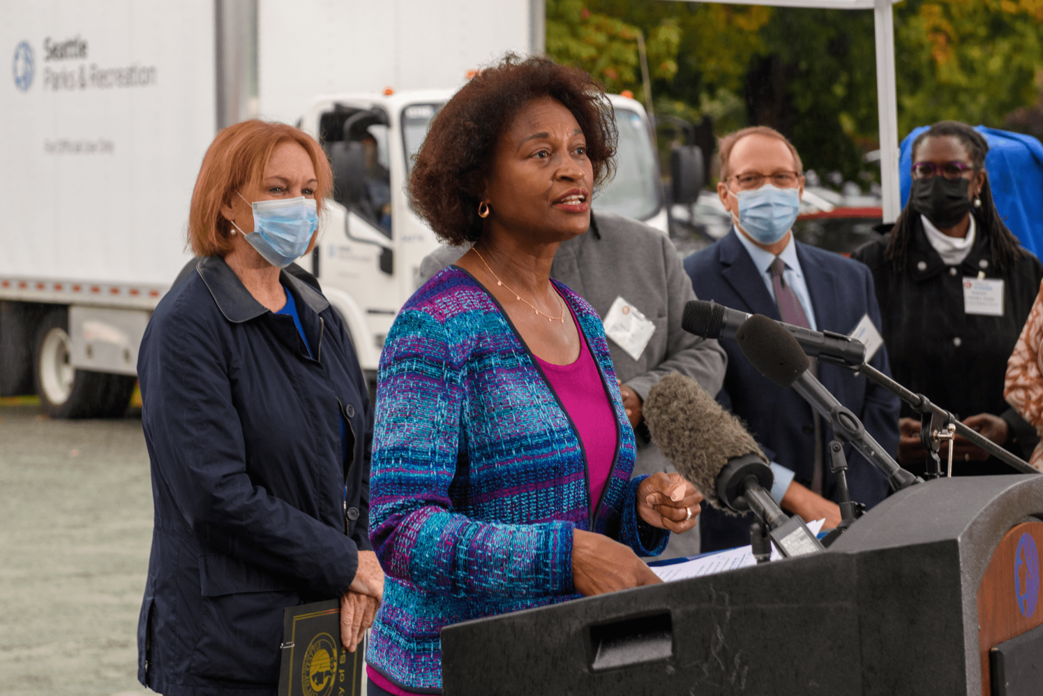 Nuria Fernandez, Administrator of the Federal Transit Administration, shares remarks with the media at a podium during a groundbreaking event in Seattle. Additional agency representatives and elected officials are visible behind her, to the left and right sides. Several microphones are visible in the foreground, and a white truck is visible in the background.