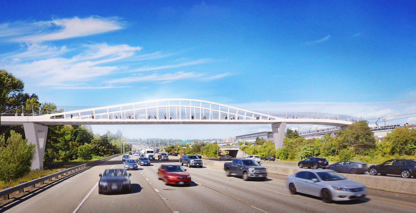 View of the new John Lewis Memorial Bridge design, looking north from I-5. Cars and trucks are visible driving by in both directions, in the foreground. A visualization of the new elevated pedestrian and bicycle bridge is visible in the background, in the center of the image. Mostly clear blue sky shine down from above.