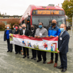 Photo of elected officials and agency representatives holding a banner in front of a bus during a groundbreaking event for the start of the Madison - RapidRide G Line project in Seattle. Nine people are standing side by side in a gravel parking lot celebrating the start of construction.