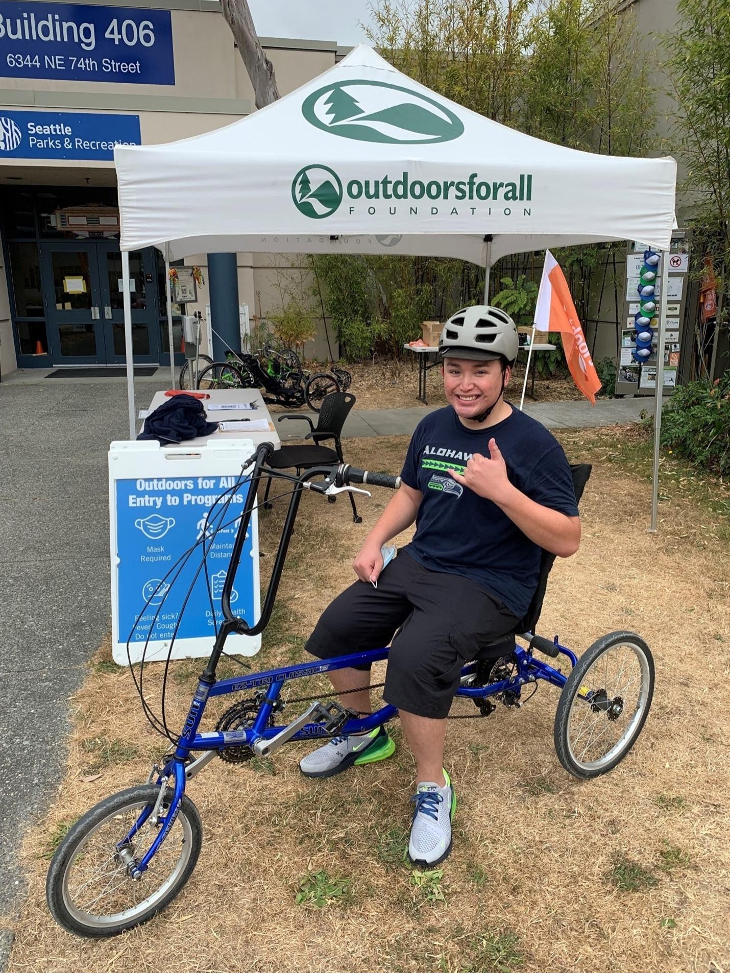 Austin smiles and gives a hang loose sign while enjoying an adaptive cycle at Magnuson Park.