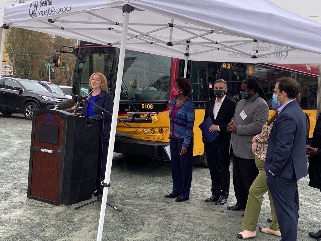 Mayor Jenny A. Durkan shares her remarks with members of the media at a groundbreaking event in Seattle. She stands at a podium and speaks into microphones. Behind her a bus is visible, and several other representatives are standing to her right.