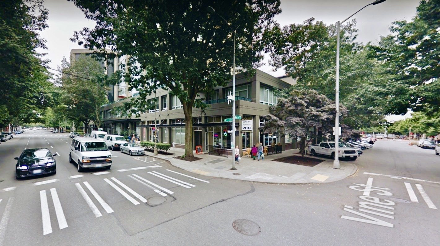 Photo of the intersection of 4th Ave and Vine St in Seattle's Belltown neighborhood. To the left a car and a van await a green light. A restaurant in visible in the middle of the photo. Many trees and several light poles are visible throughout the photo. Pedestrians walk on the sidewalk in the middle of the photo.