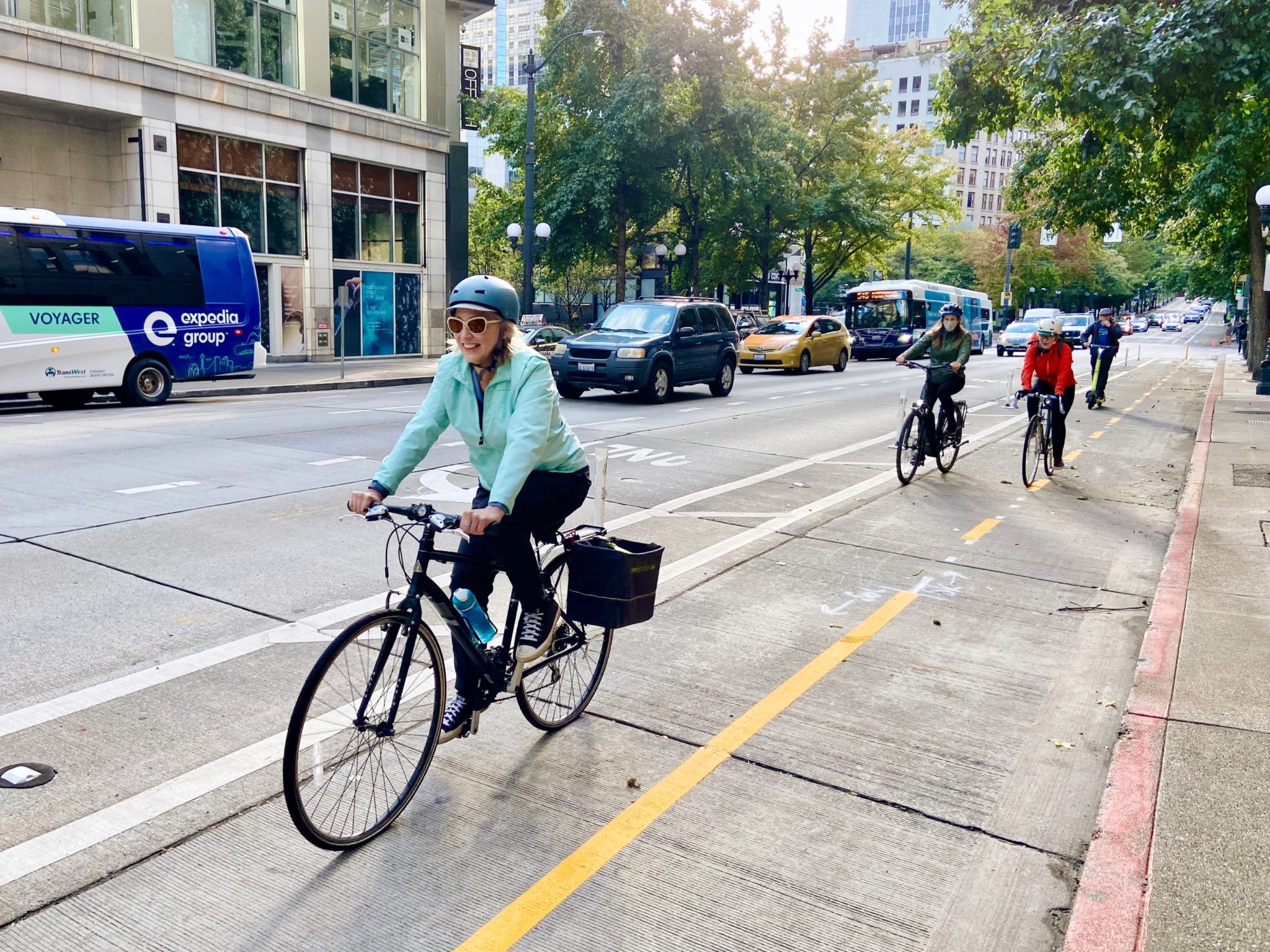 Several people bike along a protected bike lane with buses and cars traveling along the street next to them, in the same direction. Buildings and trees are in the background.