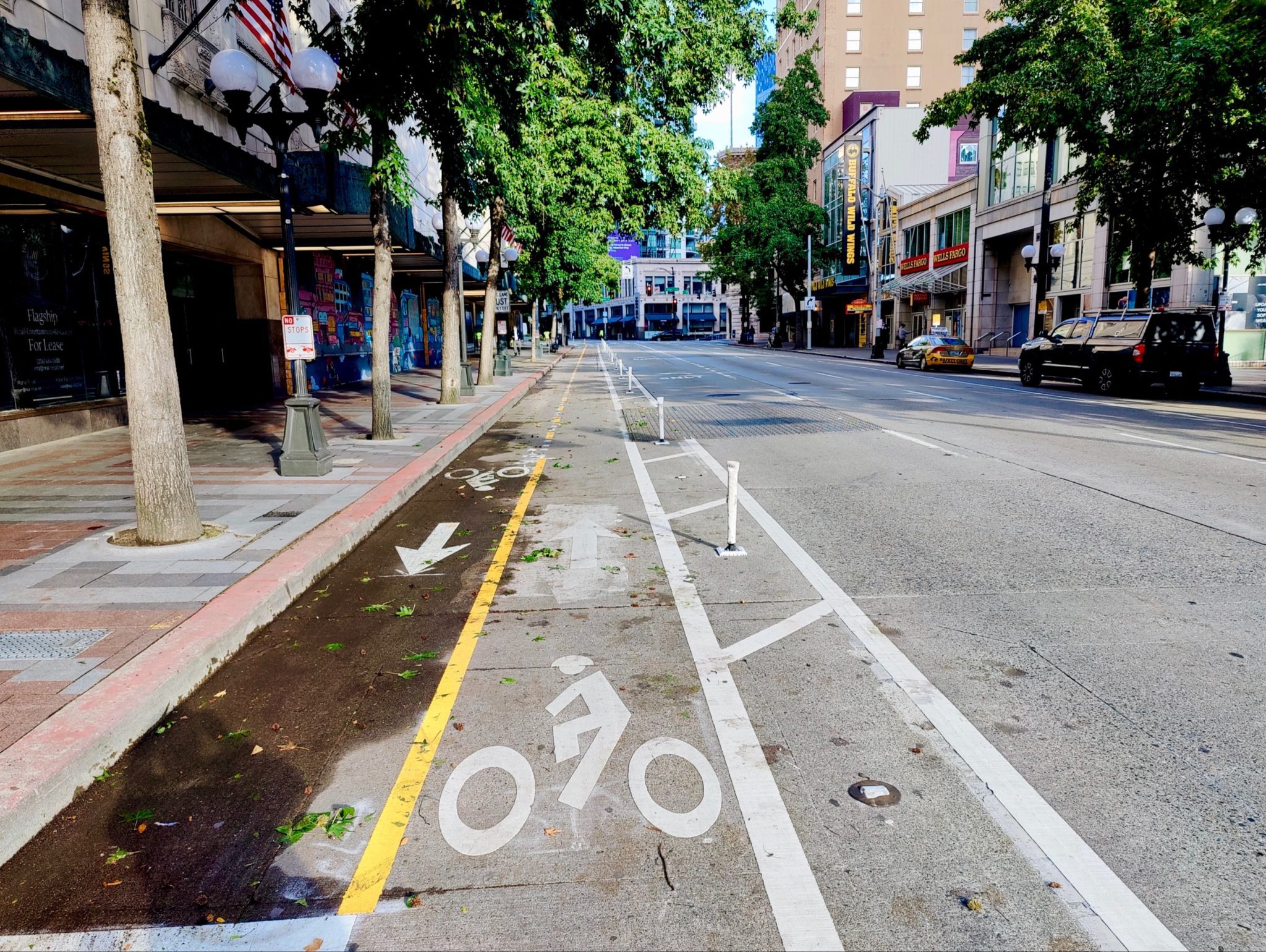 Photo of the new 4th Ave protected bike lane in downtown Seattle. The new two-wake bike lane is visible in the center, extending into the background. The sidewalk is visible on the left, and vehicular lanes of 4th Ave to the right. Buildings and trees are also visible in the photo.