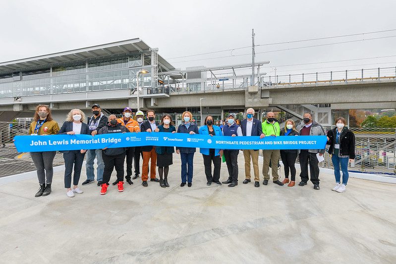 Local elected officials and agency leaders, as well as community organization representatives, prepare to cut the ribbon for the new pedestrian and bicycle bridge in Seattle's Northgate neighborhood. The new Link light rail station is visible in the background, in the upper left corner of the photo.