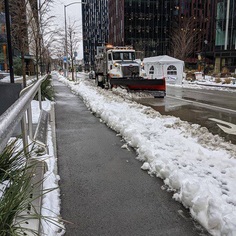 Photo of a sidewalk in the City of Seattle. The sidewalk has been cleared to allow for a smooth path for people to use, with the remaining snow off to the right of the sidewalk's path. A snowplow and large buildings are visible in the upper right side of the photo.