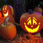 Several pumpkins and carved jack-o-lanterns sit on the ground outdoors. Two of the jack-o-lanterns are lit up by candle light, and are smiling toward the camera.