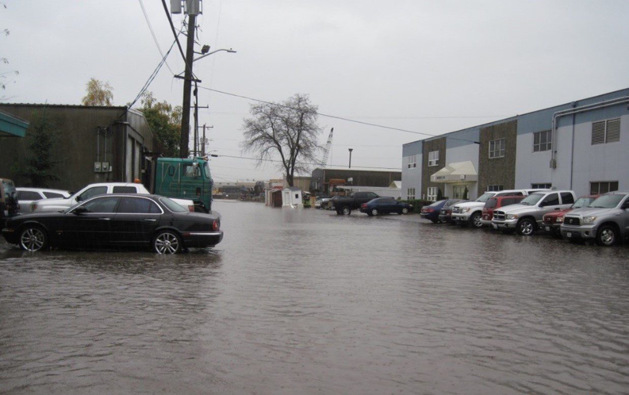 Photo of flooding during a heavy rain event. Parks cars are visible on the left and right sides, with flooded roadway in the main part of the image.