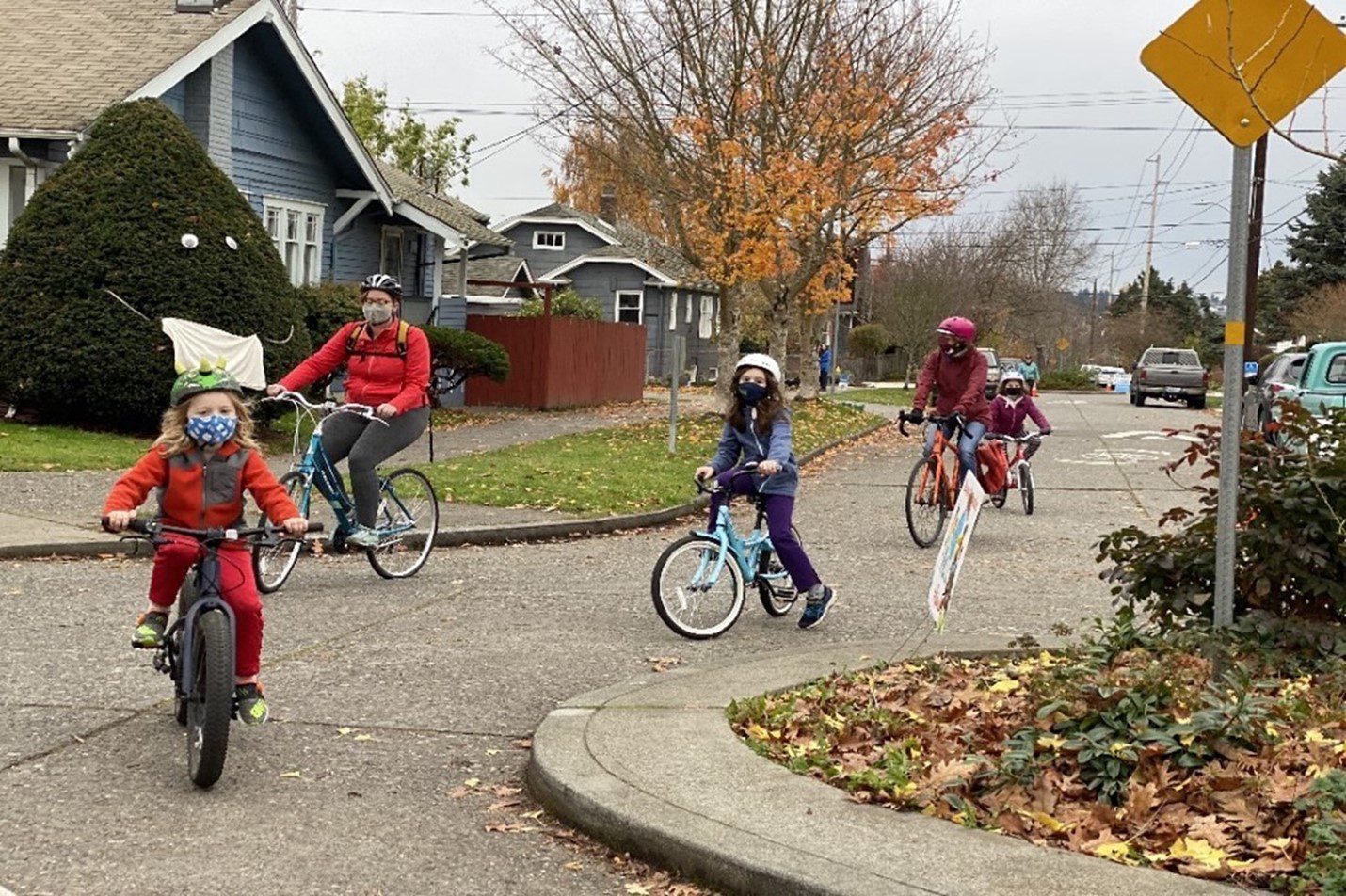 A number of school aged children ride their bicycles to school alongside two parents, on a local street in Seattle. There are a total of three children and two adults pictured, all on bicycles. A pedestrian is visible in the background. Fallen leaves and several parked cars, and nearby houses, can be seen in the foreground and background of the image.