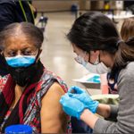 A woman receives a dose of the COVID-19 vaccine at a vaccination site in Seattle. Both the woman receiving the vaccine and the person administering it are wearing masks. The worker wears light blue medical gloves. Other people are visible in the background of the indoor site.