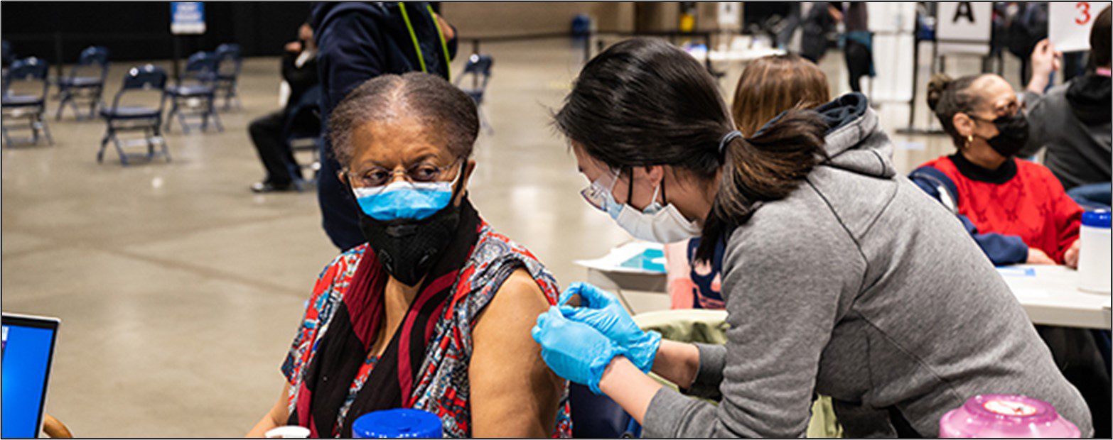 A woman receives a dose of the COVID-19 vaccine at a vaccination site in Seattle. Both the woman receiving the vaccine and the person administering it are wearing masks. The worker wears light blue medical gloves. Other people are visible in the background of the indoor site.