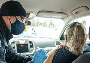 Image of a woman receiving a dose of the COVID-19 vaccine at a free vaccination location in Seattle. The man administering the dose is wearing blue gloves and a face mask. The woman is sitting in her vehicle while she receives the vaccine.