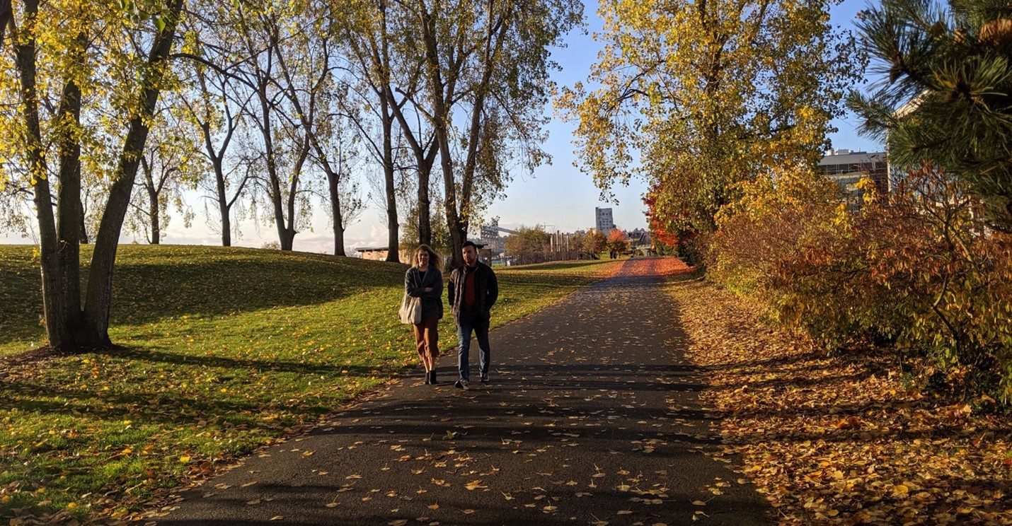 Two people walk along a path in Seattle, surrounded by trees, grass, and fallen leaves. The man and woman are in the center of the photo, with the trail extending into the background. Light blue skies are visible near the top of the image.