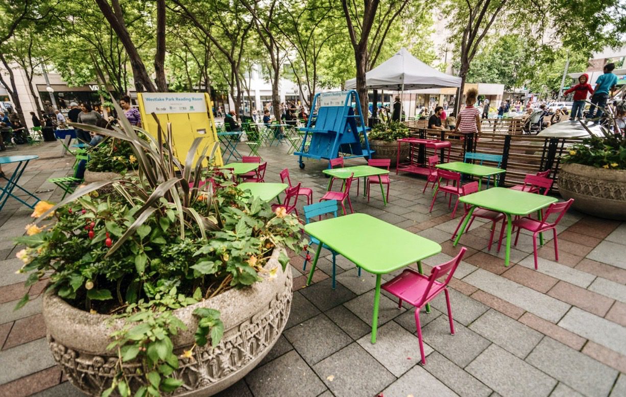 Photo of Seattle's Westlake Park plaza. Several colorful tables and chairs are visible in the foreground alongside a large cement planter box with vegetation growing inside. Children are visible playing in a kids play area to the upper right of the photo. Many trees are visible near the top of the photo.