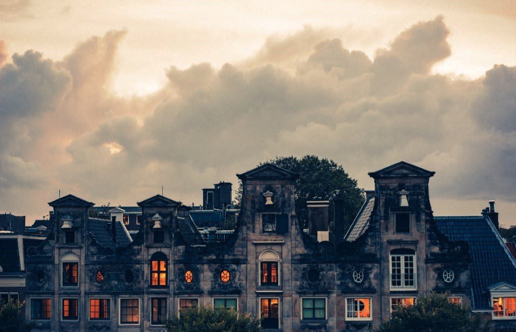 A large mansion has orange lights on in its interior rooms. Ominous gray clouds loom in the sky above.