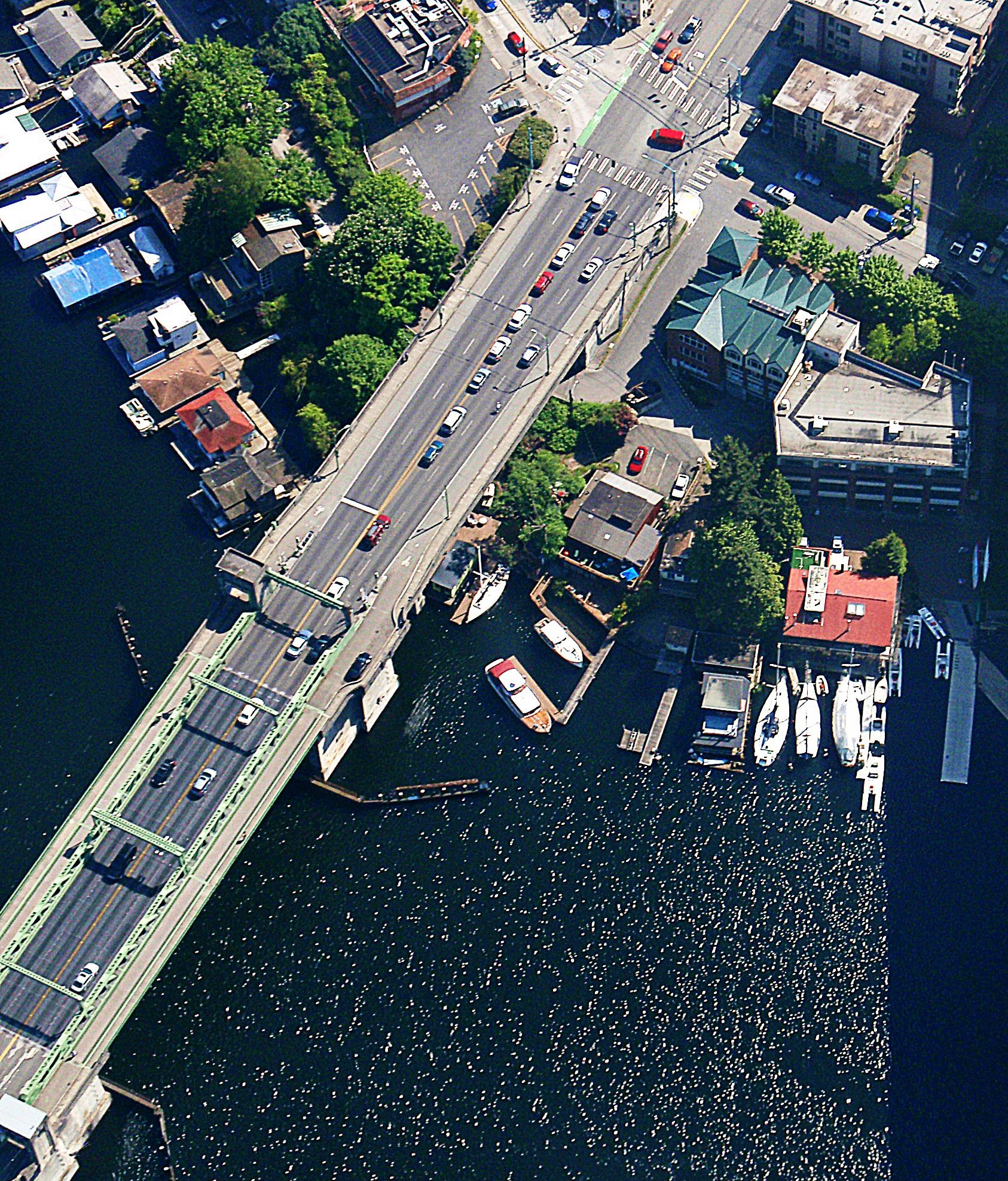 Aerial photo of the University Bridge with traffic flowing across the Ship Canal in May 2012. The bridge is visible to the lefthand side of the photo, with a number of boats and buildings visible near the top. Sunlight reflects back off of the water in the lower righthand side of the image.
