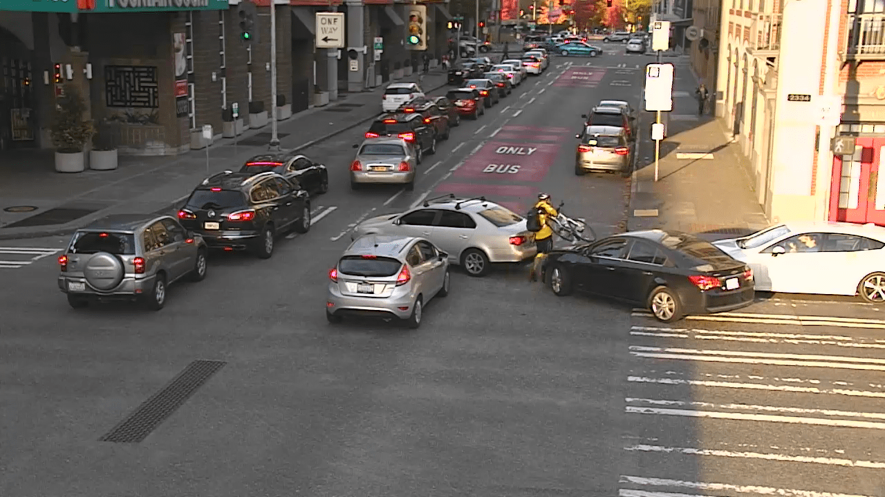 Several vehicles block an interersection, crosswalk, and bus only lane in Seattle's Belltown neighborhood. Numerous cars are stuck in a traffic jam, and a person biking must pick up their bicycle and walk it through traffic while navigating the blocked crosswalk. Buildings are visible on either side of the street and the crosswalk on the righthand side.